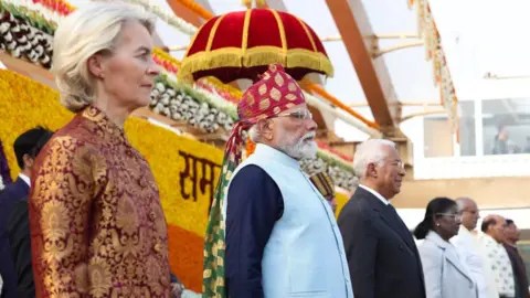 Narendra Modi/X Leaders stand in a line on a decorated stage at an outdoor ceremony. From left to right are a European woman leader in a red patterned jacket, Indian Prime Minister Narendra Modi wearing a traditional turban and light blue vest, a European male leader in a dark suit, and Indian President Droupadi Murmu in a light-coloured coat, with floral decorations and officials visible in the background