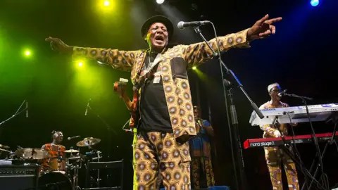 Getty Images Ebo Taylor in a yellow African-print suit on stage. He is singing and has his arms stretched out either side of him.