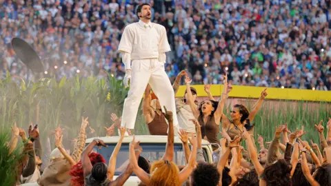 Reuters Bad Bunny on stage at the Superbowl. He is standing on a car and surrounded by female dancers waving their arms. 