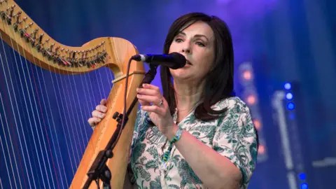 Getty Moya Brennan playing a harp and singing. She is in a green and white dress.