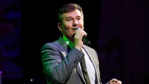 Getty Daniel O'Donnell is wearing a grey suit jacket, a white shirt and a black tie as he sings into a microphone. He is performing on stage, with a green light just behind him reflecting off his brown, short hair.