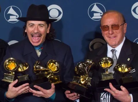 Getty Images Clive Davis and Santana celebrate their success at the 2000 Grammy Awards, they are both wearing black suits and holding several Grammy trophies each.